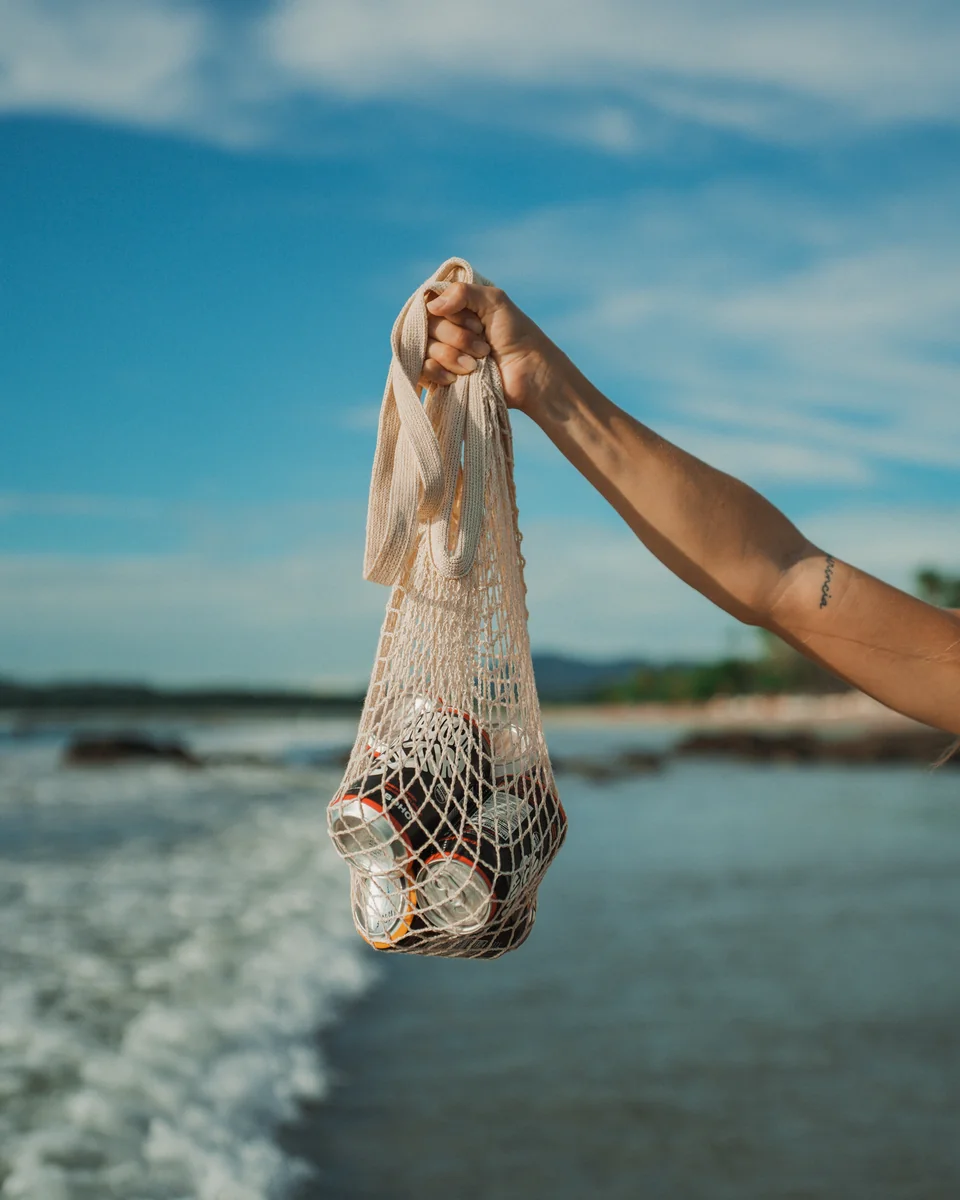 Hand holding a mesh beach tote filled with canned drinks against the ocean and blue sky in Guanacaste, Costa Rica