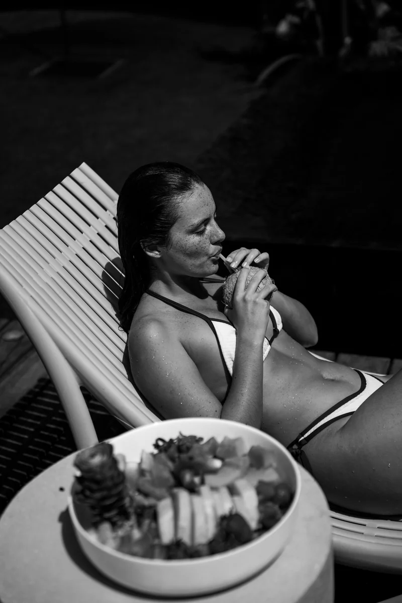 Black-and-white photo of a woman sipping from a coconut on a poolside lounger beside a fruit bowl, lifestyle photography in Costa Rica