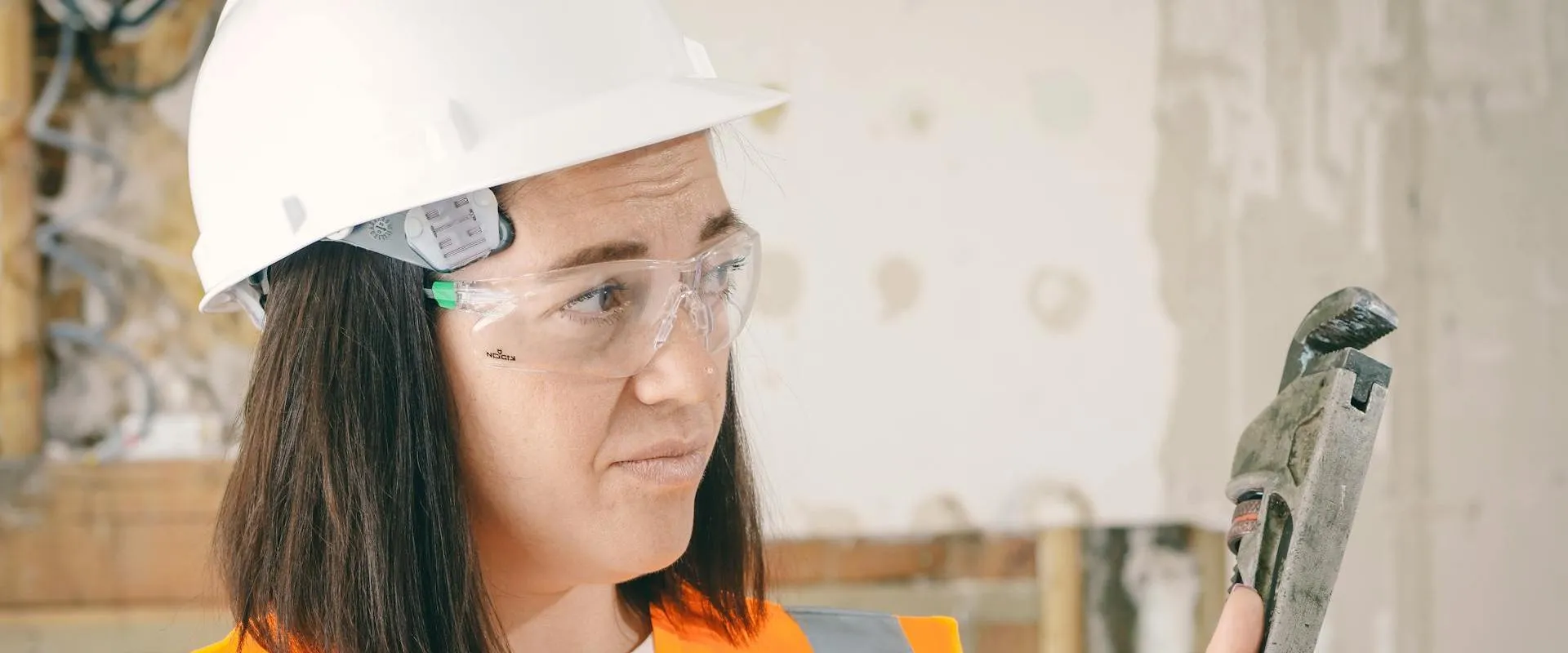 A woman wearing safety equipment holds a plumbing wrench.