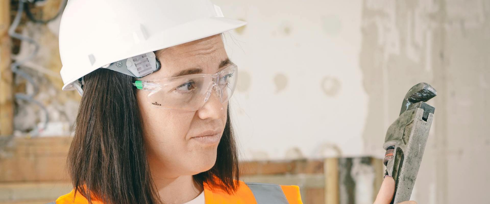 A woman wearing safety equipment holds a plumbing wrench.