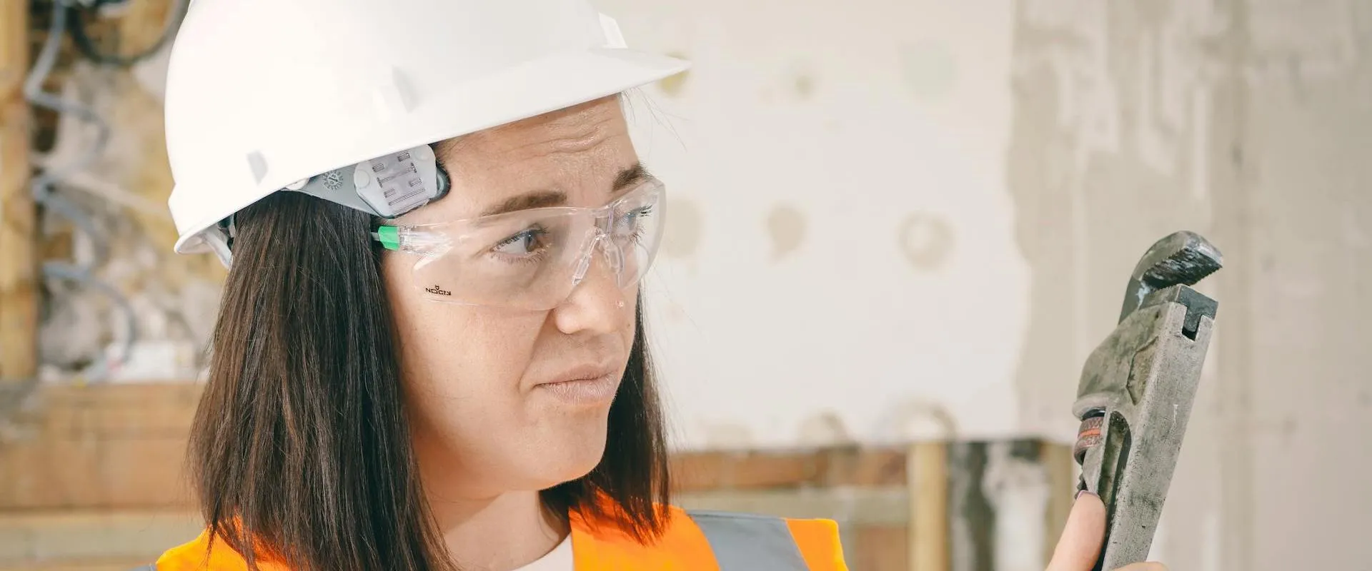 A woman wearing safety equipment holds a plumbing wrench.