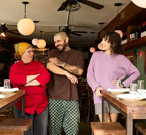 Three people stand side by side, smiling in a cozy restaurant. The atmosphere is warm and friendly, with wooden tables, chairs, and soft lighting.