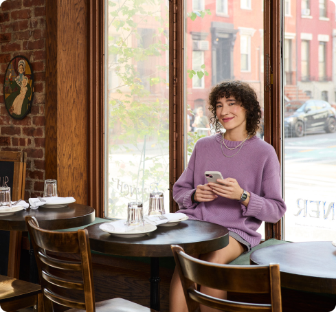 A woman in a purple sweater sits at a round table in a cozy cafe, smiling at her phone. Large windows reveal a street with red brick buildings.