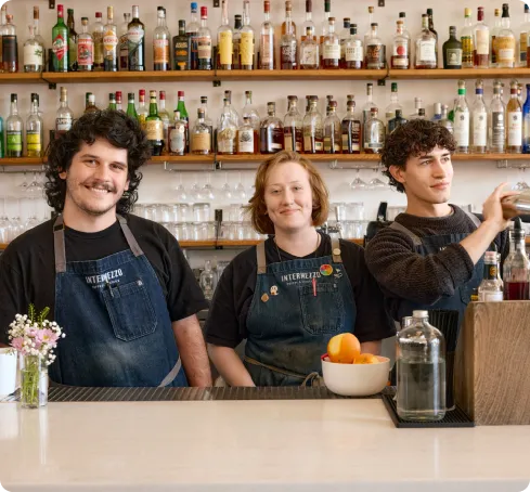 Three smiling bartenders stand behind a bar, wearing aprons. Shelves of colorful liquor bottles line the wall. A vase of flowers adds a cheerful touch.