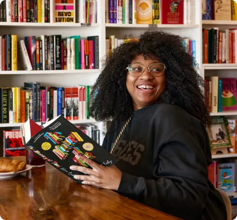 Smiling woman with glasses reads a colorful book at a wooden table in a cozy bookstore. Bookshelves filled with various titles in the background.
