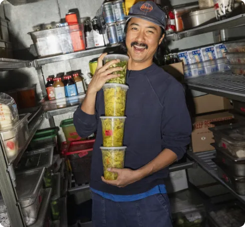 A person is smiling broadly in a restaurant storage room, holding a stack of containers filled with green pickled vegetables. Shelves with various food items surround them.