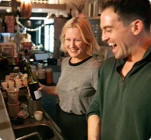 Two smiling staff members laugh together behind a bar counter, with one holding a wine bottle in a warmly lit restaurant or café setting.