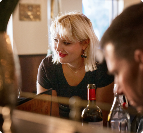 A smiling server with short blonde hair leans toward a customer across a bar or restaurant counter, with wine bottles visible in the foreground.