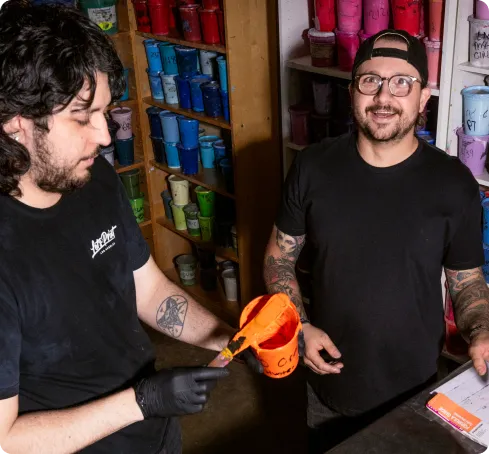 Two tattooed workers in black t-shirts collaborate in a paint or craft shop, one painting an orange bucket while the other looks on, with colorful cups lining the shelves behind them.