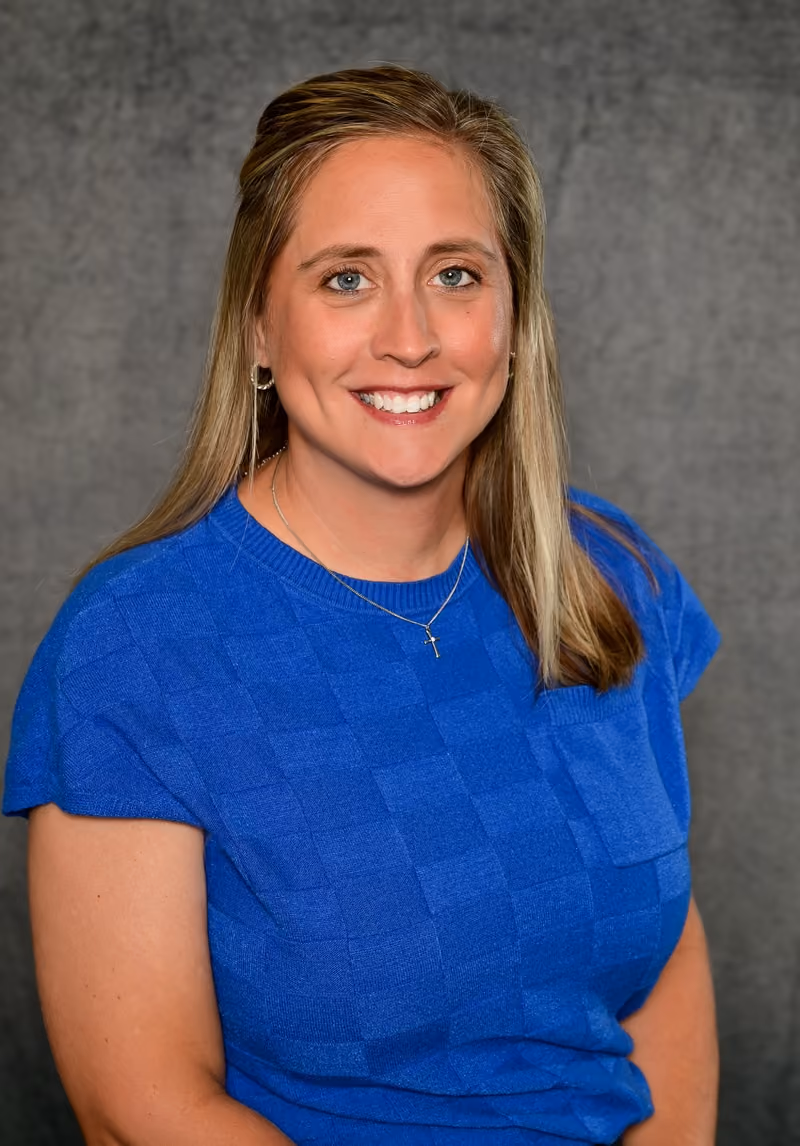 Smiling woman with blonde hair wearing a blue textured short-sleeve shirt and a cross necklace.