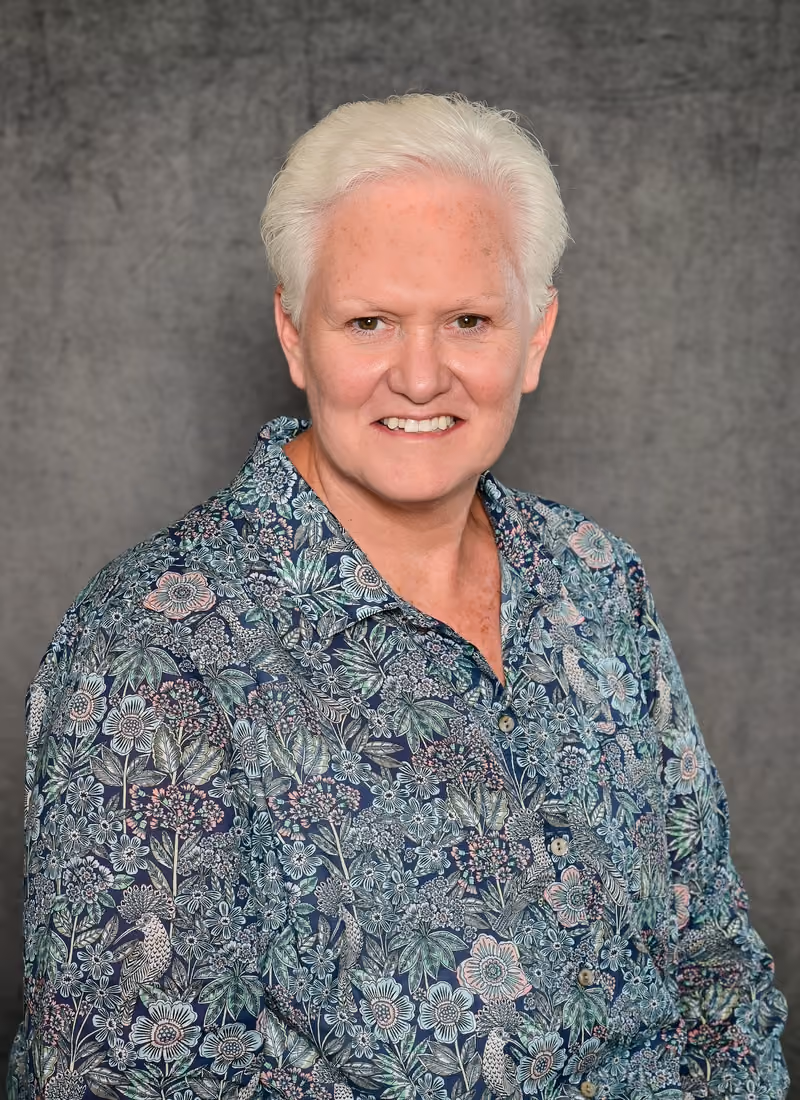 Smiling older woman with short white hair wearing a blue floral patterned shirt against gray background.