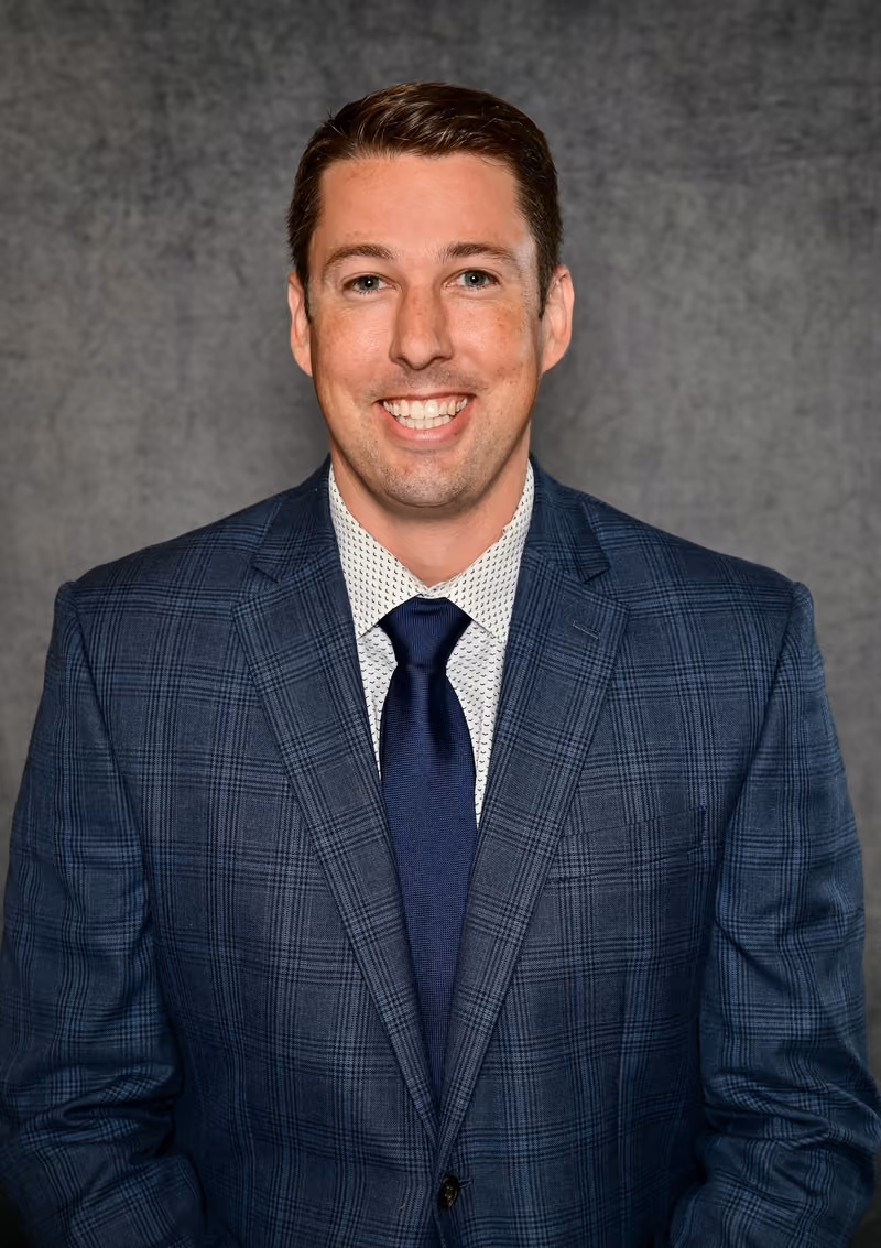 Smiling man wearing a blue plaid suit, white patterned shirt, and navy tie, posing against gray background.