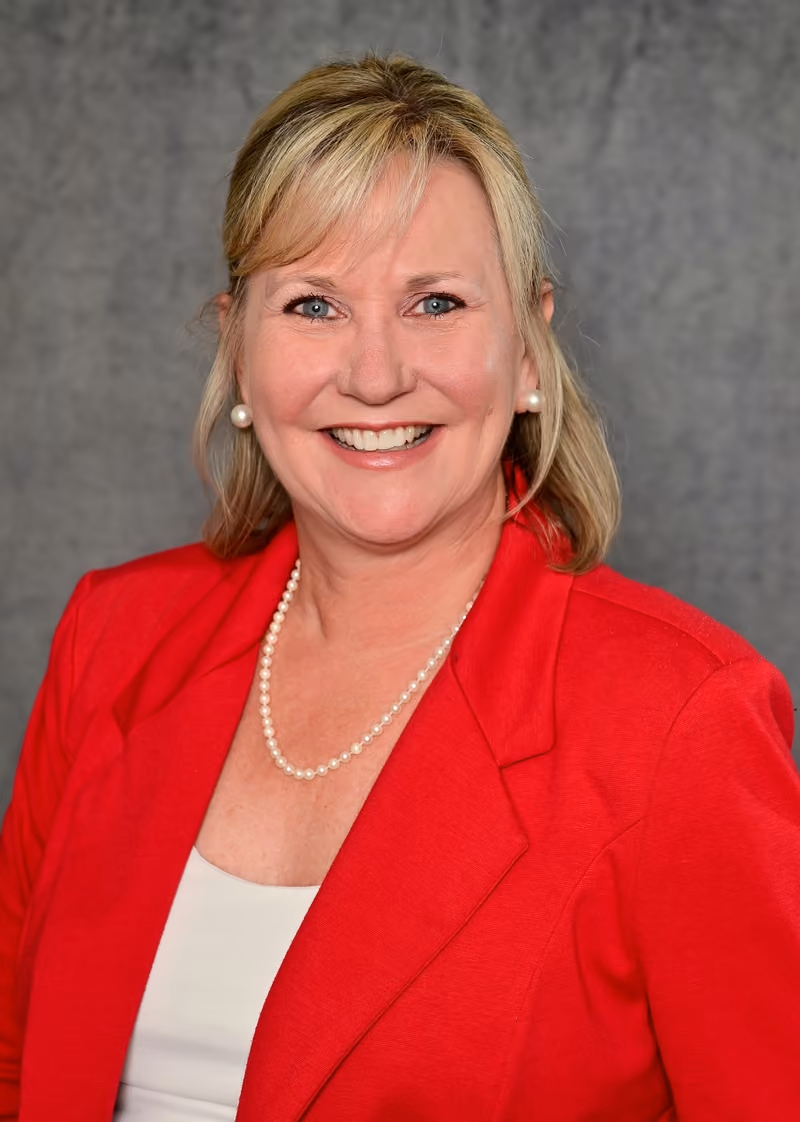 Smiling blonde woman wearing pearl earrings, a pearl necklace, white top, and red blazer against gray background.