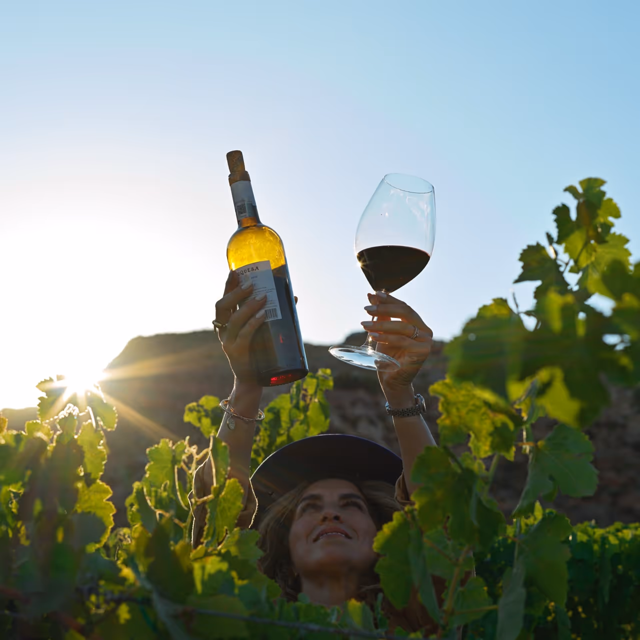Una mujer sosteniendo una botella de vino y una copa, en medio de un viñedo. Rutas Enoturísticas de Baja California