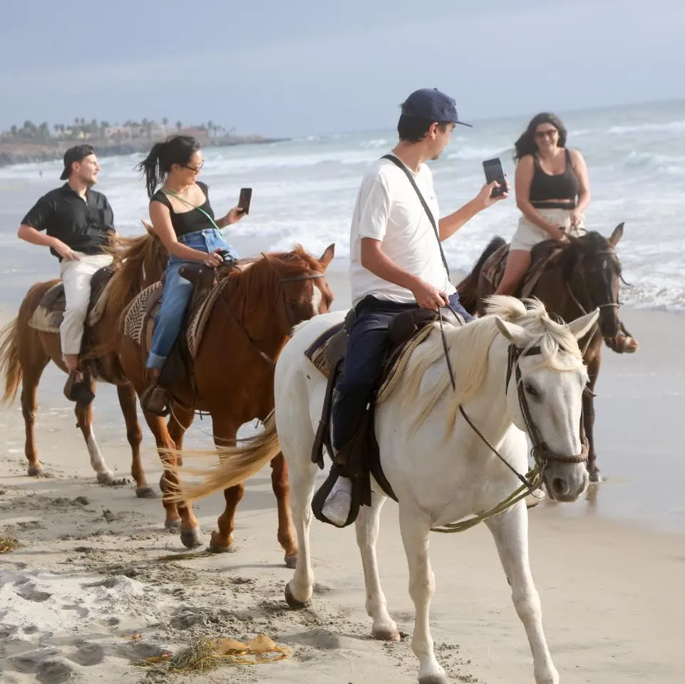 Turistas cabalgando frente al mar, mientras toman fotos. Una cabalgata grupal en La Ruta Vaquera, Baja California