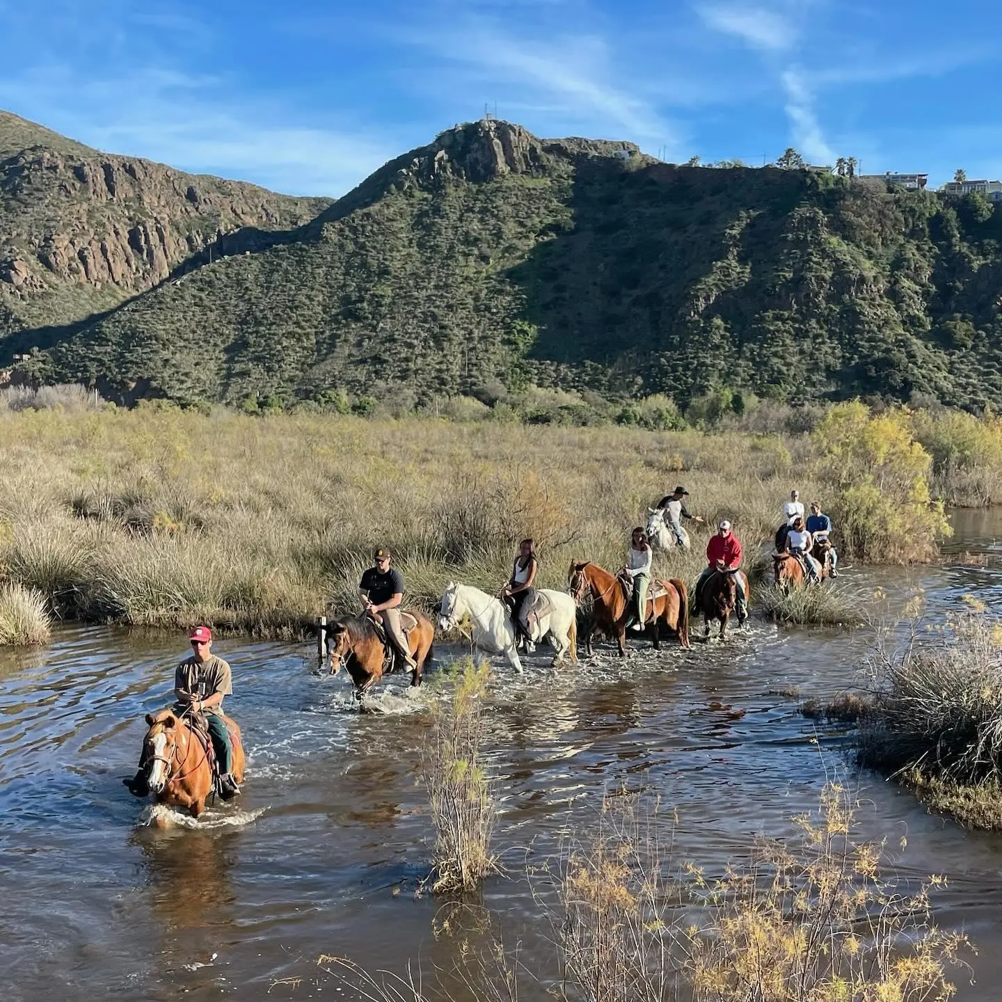 UN grupo de personas cabalgando atravesando un pequeño cuerpo de agua entre montañas. Una festividad en ensenada con humo , luces y mucha gente disfrutando a caballo en Ensenada. La Ruta Vaquera, Baja California