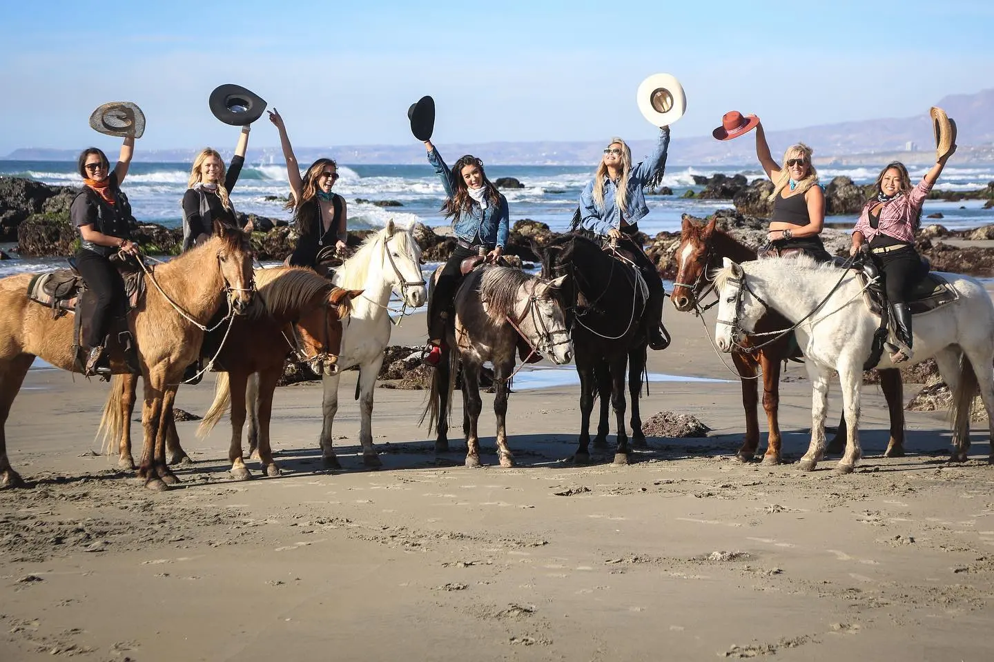 Un grupo de turistas posando para una foto, levantando un sombrero vaquero con su mano. En medio de la playa, con las rocas, el mar y el cielo fe fondo. La Ruta Vaquera, Baja California