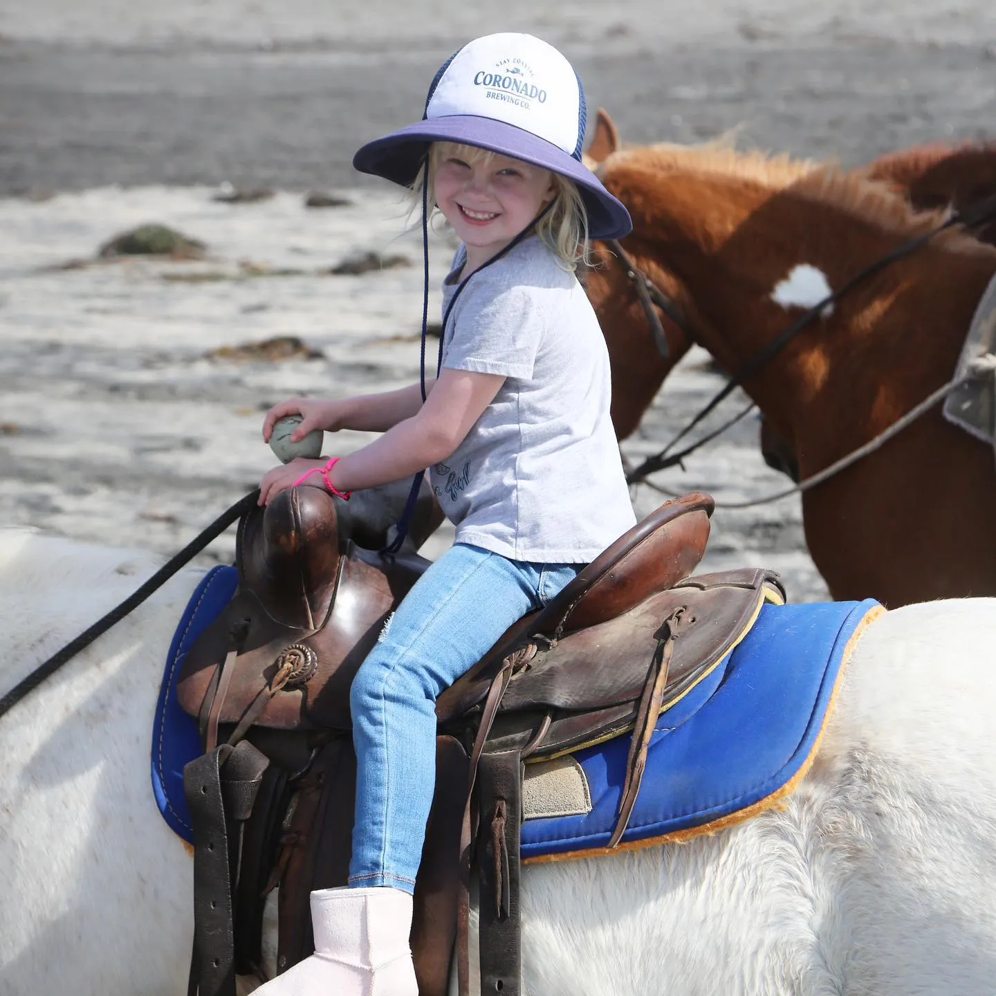 Una niña pequeña montando a caballo y sonriendo a la cámara con la playa y las olas de fondo. Una festividad en ensenada con humo , luces y mucha gente disfrutando a caballo en Ensenada. La Ruta Vaquera, Baja California