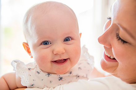 A baby smiling at the camera while in the arms of their mother