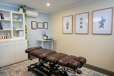 Therapy room with adjustable brown leather treatment table, white cabinet, wall art, and a small table with a plant.