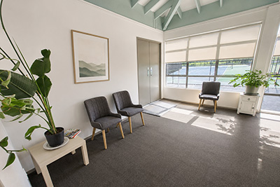 Waiting room with three gray chairs, plants, a small table, and large windows with blinds.