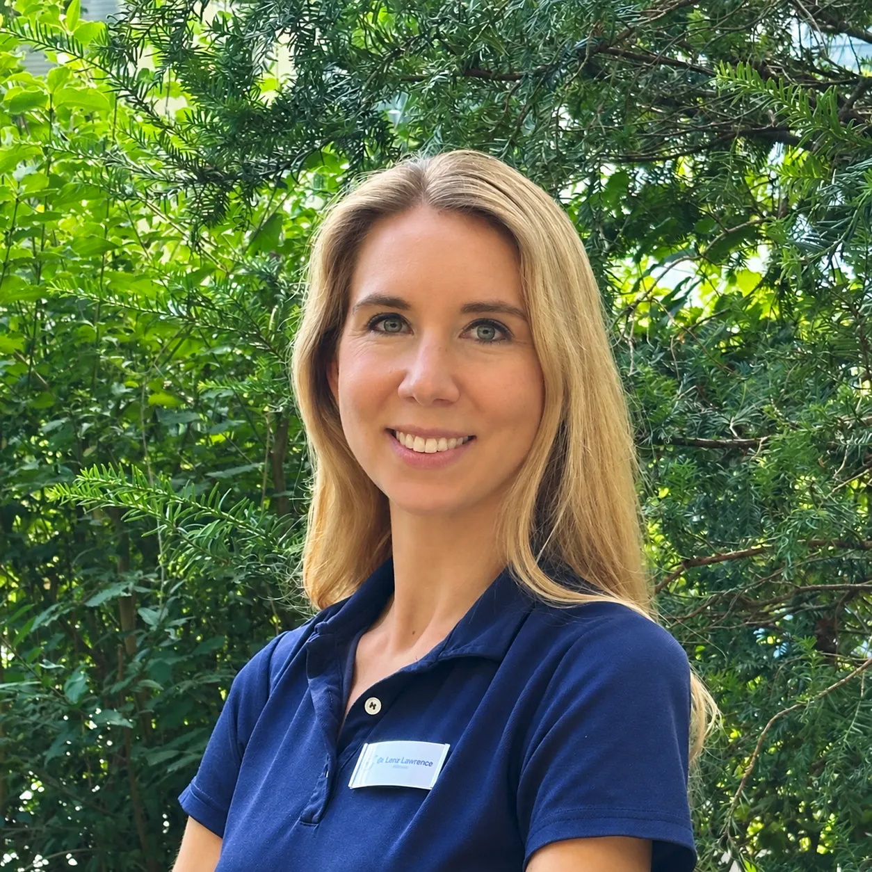Eine lächelnde Frau mit langen blonden Haaren, die ein dunkelblaues Poloshirt und ein Namensschild trägt, steht im Freien vor grünem Laub.
