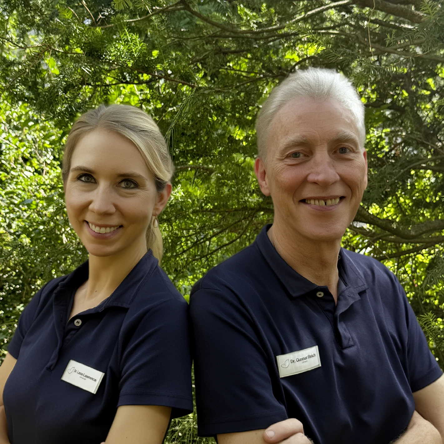 Eine lächelnde Frau mit langen blonden Haaren, die ein dunkelblaues Poloshirt und ein Namensschild trägt, und ein lächelnder grauhaariger Mann mit dunkelblauem Polo und Namensschild stehen im Freien vor grünem Laub.