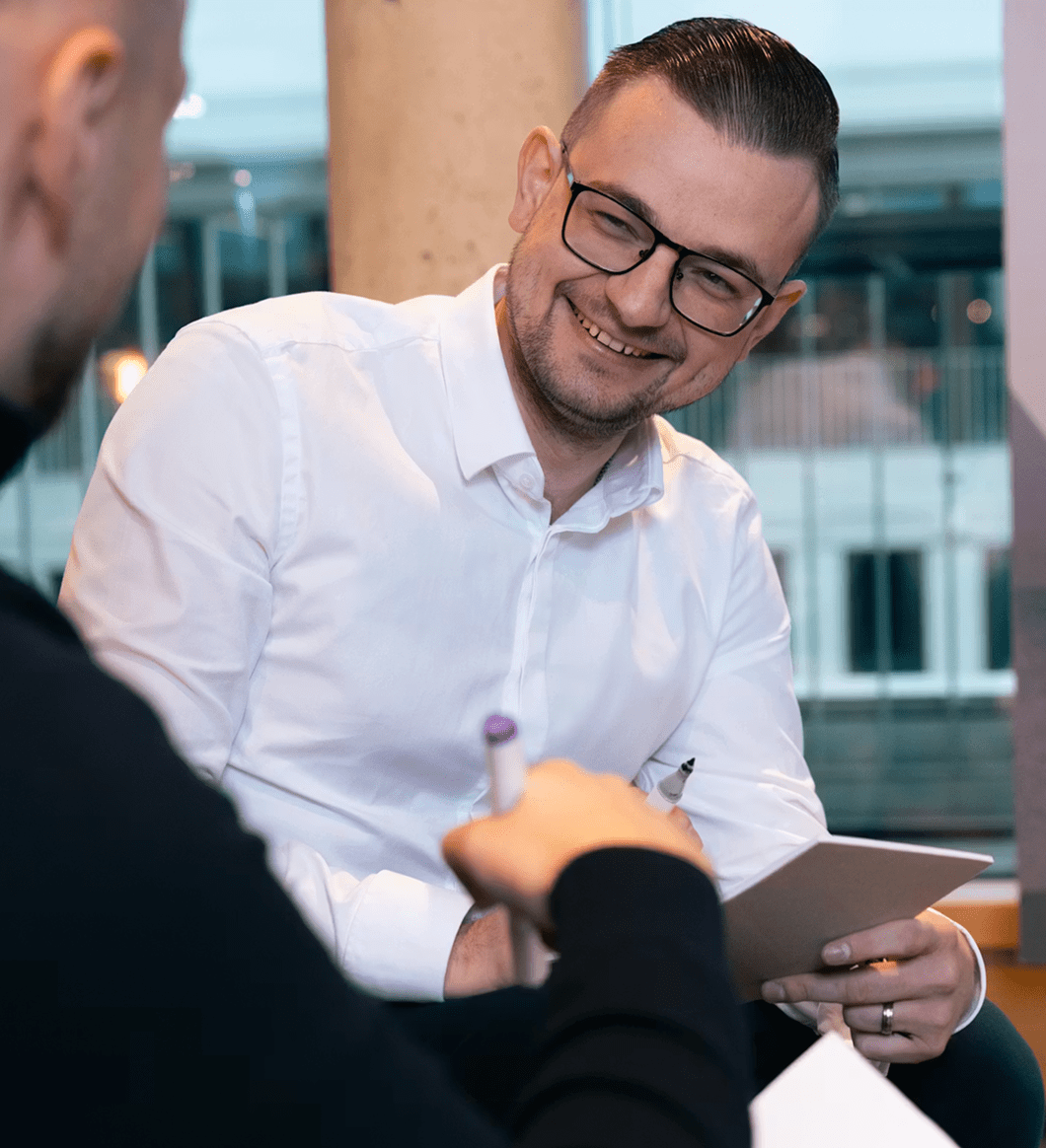 Ein Mann mit Brille und weißem Hemd sitzt in einer modernen Umgebung und lächelt im Gespräch. Er hält ein Tablet in der Hand, während ihm eine zweite Person mit Stift gegenübersitzt. Die Szene wirkt entspannt und kollegial.