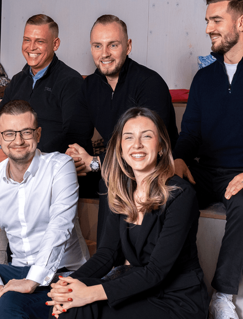 Sechs Personen sitzen locker auf Stufen und lachen gemeinsam in die Kamera. Die Szene wirkt freundlich und ungezwungen, mit moderner Büro-Atmosphäre im Hintergrund. Die Gruppe wirkt motiviert und gut gelaunt, als würde sie Teil eines eingespielten Teams sein.