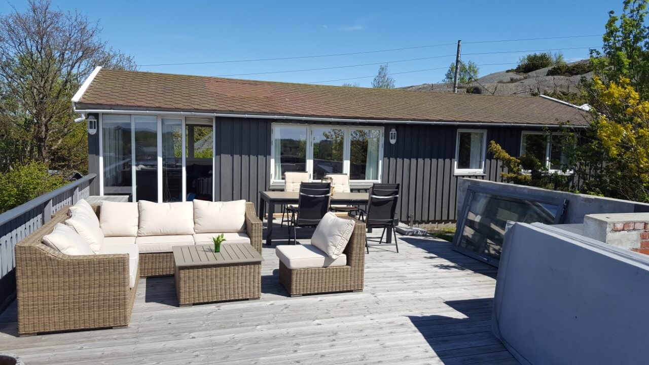 Bilde av Gunnar Eckbos hytte Sandø ved Hvasser tatt utenfra der man ser hytta med store vinduer og en lys sofa på terrassen