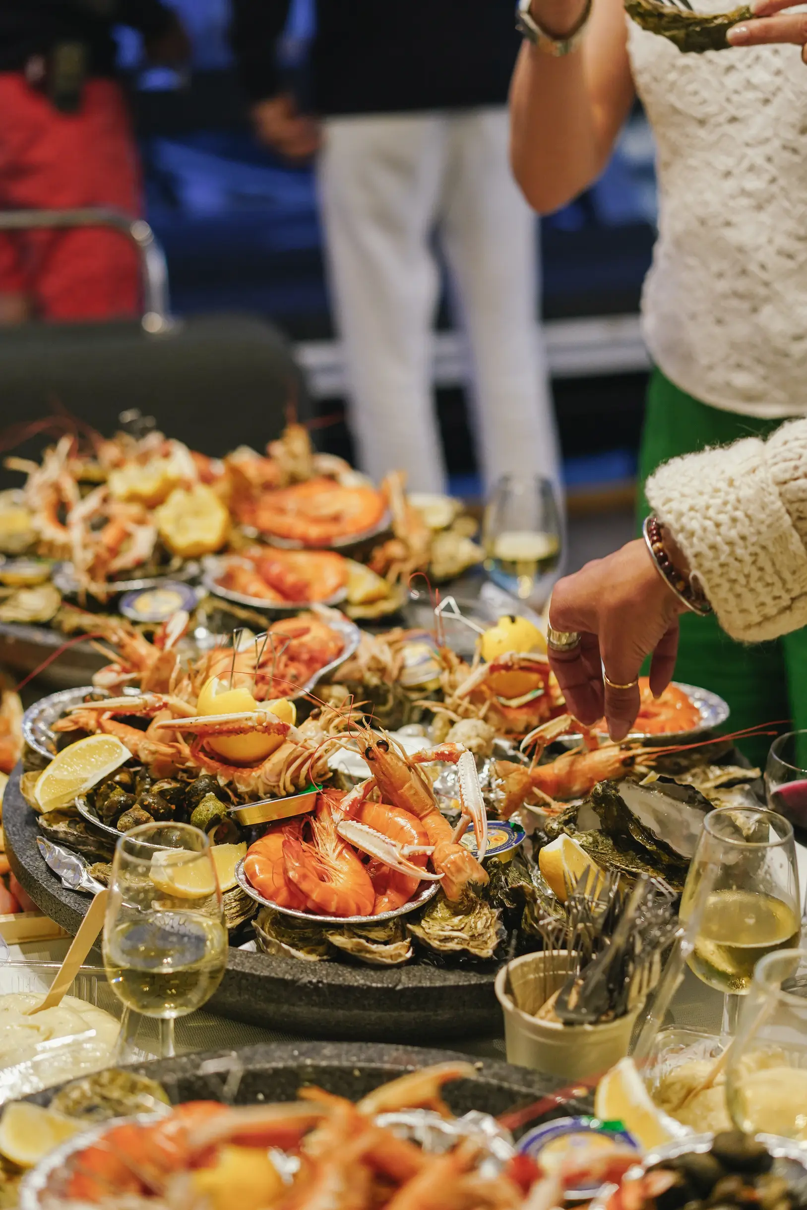 Platter of assorted seafood including shrimp, oysters, and lemon wedges, with a person reaching for food, and glasses of white wine on the table.