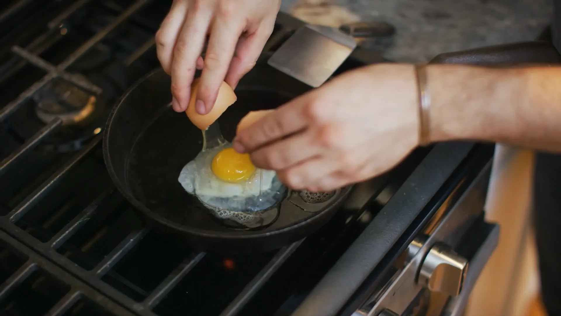 Close-up of hands cracking an egg into a hot skillet on a gas stove, with soft kitchen lighting and a casual home setting.