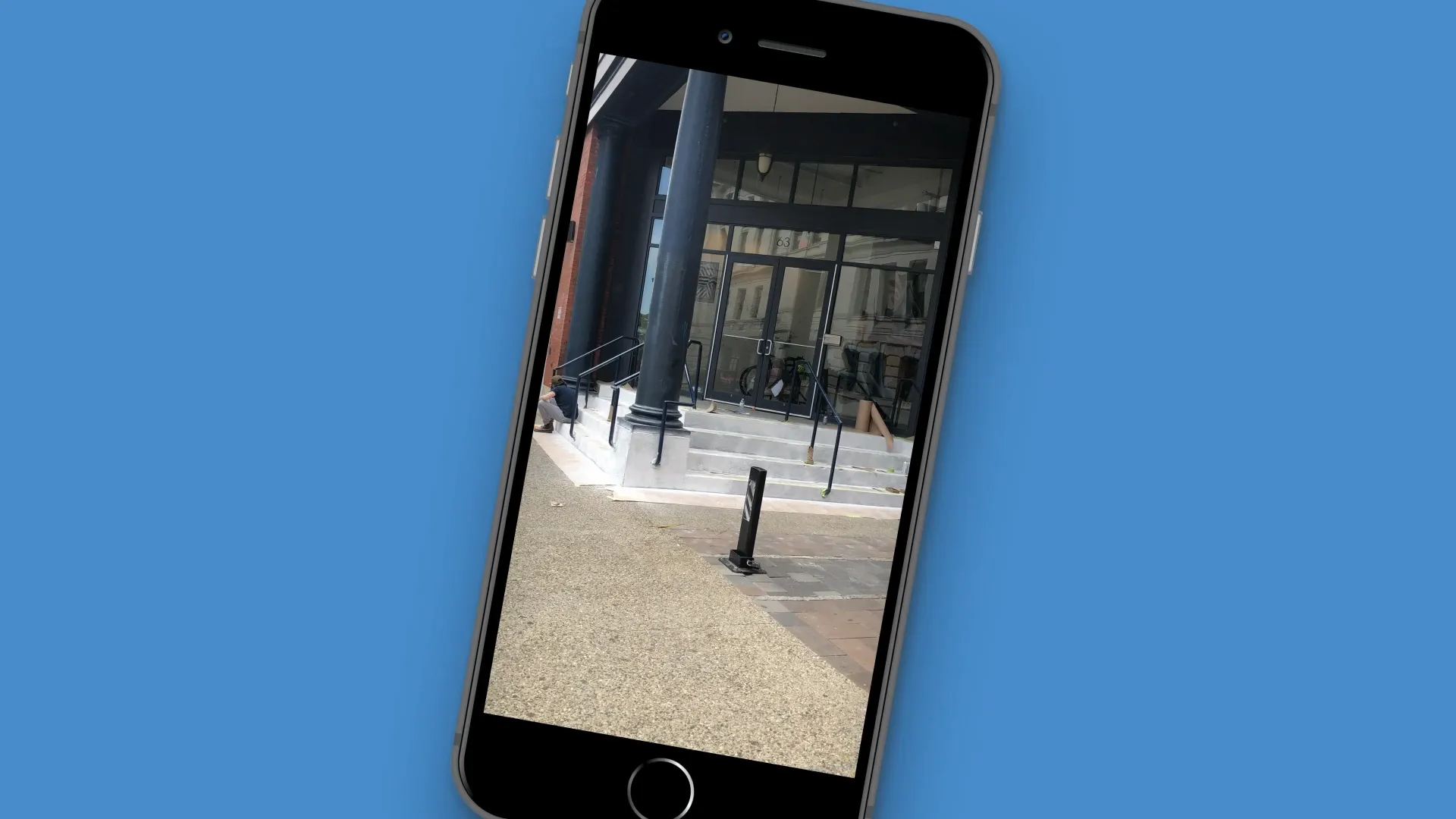 Smartphone mockup displaying photo of a modern building entrance with glass doors and black pillars, set against a solid blue background.