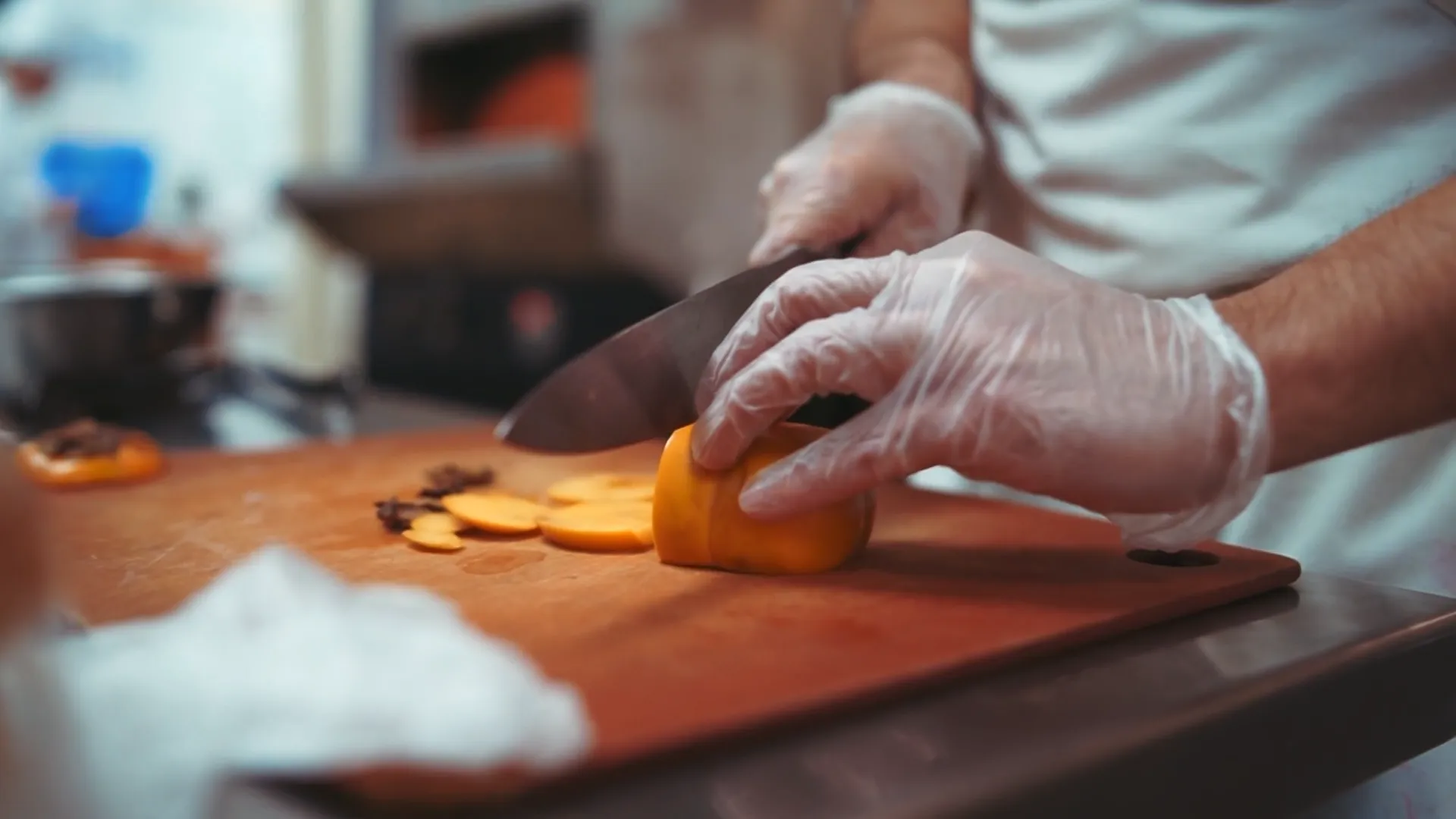 Close-up of a chef wearing gloves slicing a yellow bell pepper on a wooden cutting board in a professional kitchen setting.