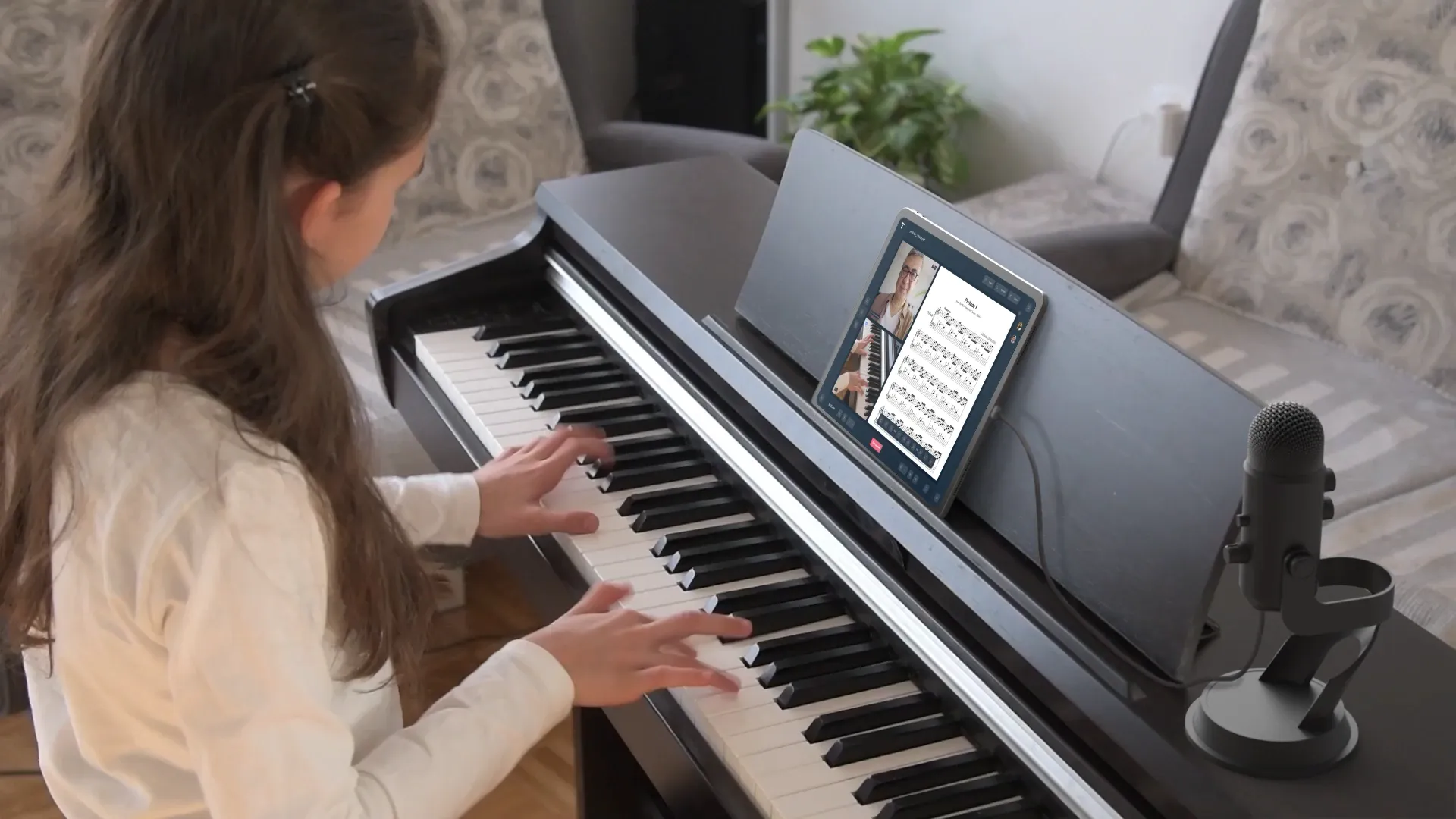 Student playing piano during an online music lesson with instructor visible on tablet screen, microphone setup beside digital keyboard.