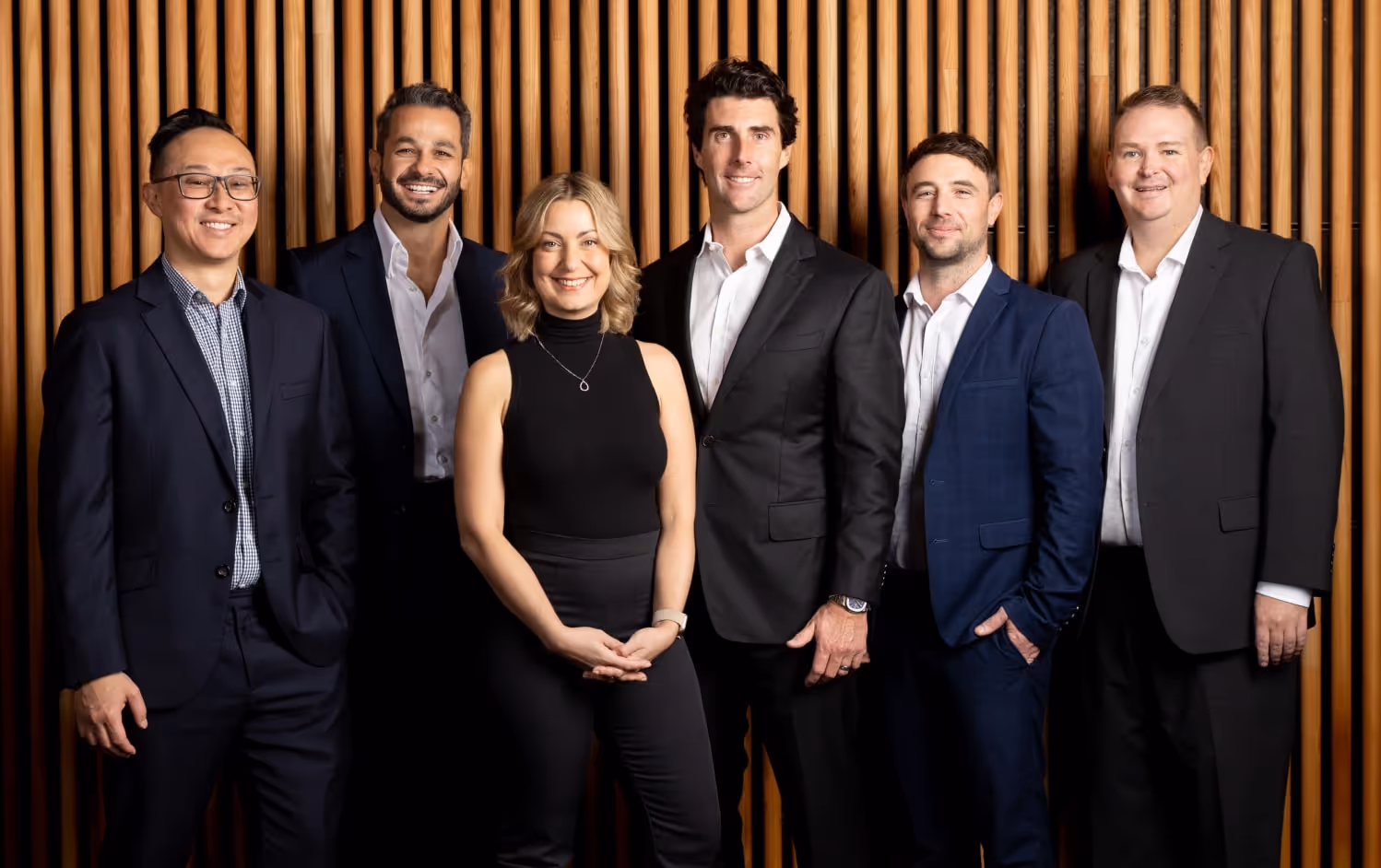 This image shows a group of six people, three men and one woman standing in a row, all dressed in business attire. They are posed in front of a wooden slat background. The group is smiling, conveying a professional yet approachable demeanor, likely representing a business team or executives.