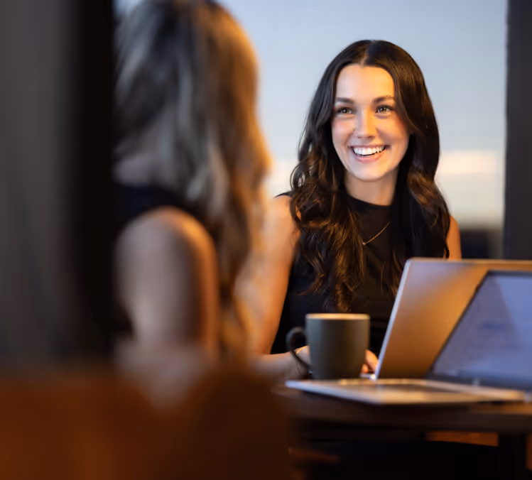 This image features a woman with long, dark hair, smiling and engaging in conversation with another person who is slightly out of focus in the foreground. She is seated at a table with a laptop and a coffee cup, suggesting a casual and collaborative work environment. The background is softly blurred, emphasizing the warmth and friendliness of the interaction.