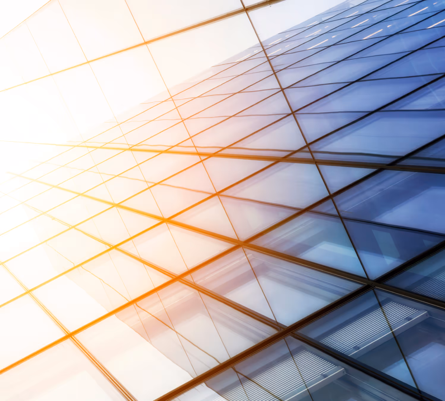 This image captures an abstract view of a modern glass building, with the focus on the reflective windows creating a geometric pattern. The photo is taken at an angle, emphasizing the lines and symmetry of the architecture. A warm sunlight glow is visible on the left side, adding a gradient of golden hues to the otherwise cool-toned glass, giving the image a dynamic and modern feel.