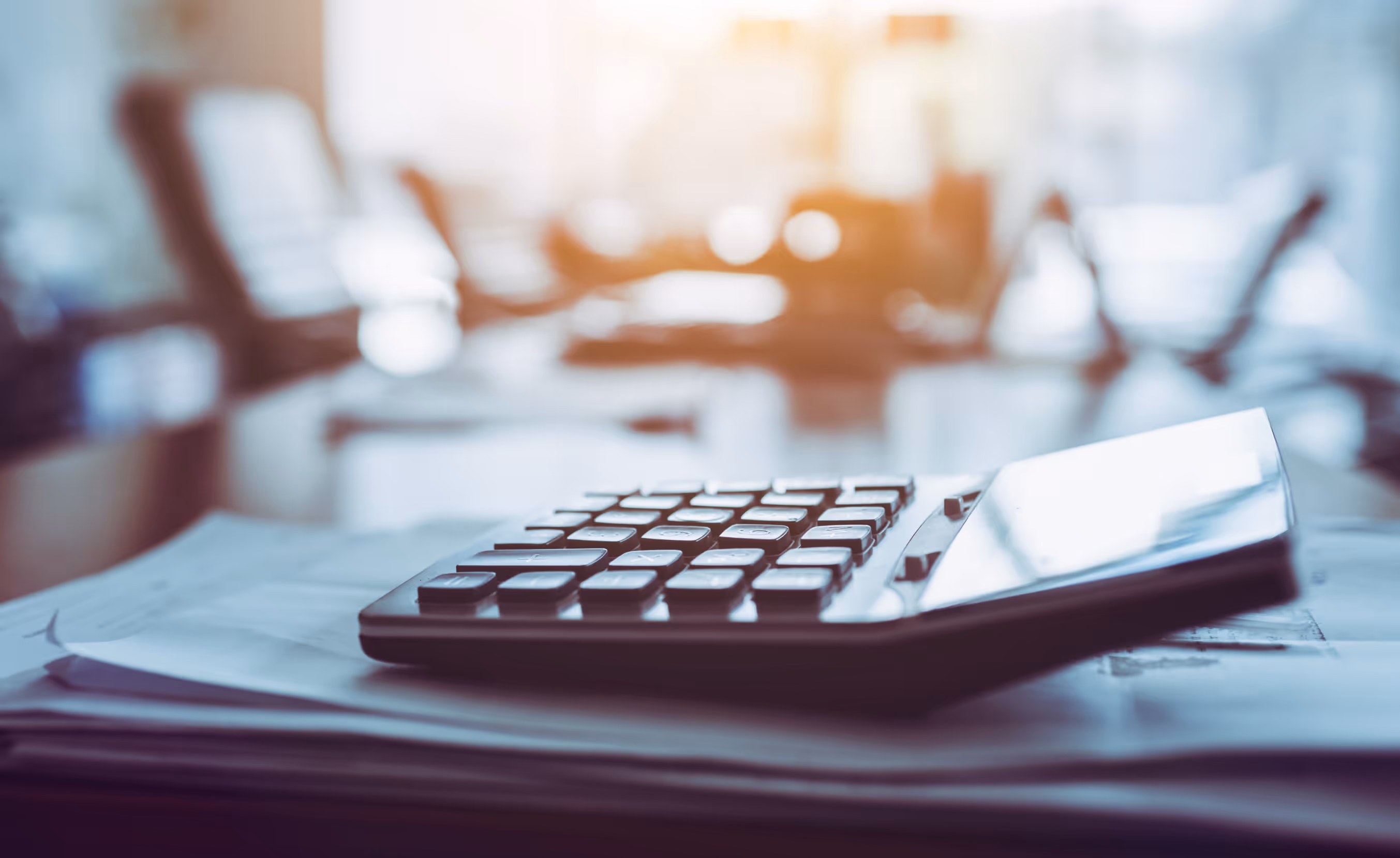 This image shows a close-up of a calculator resting on a stack of papers, likely in an office setting. The background is softly blurred, with a warm light filtering through, creating a calm and focused atmosphere. The image suggests a financial or business context, with the calculator symbolizing tasks such as budgeting, accounting, or calculating expenses.