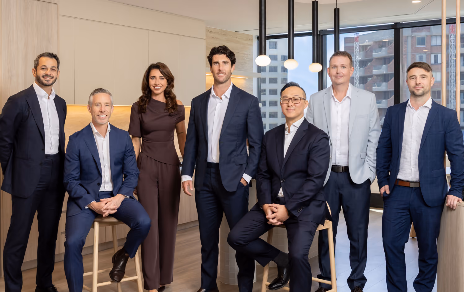 Seven professionally dressed people posing in a modern office kitchen with wooden cabinets and large windows; five men in suits and one woman in a brown outfit stand while two men sit on wooden stools.