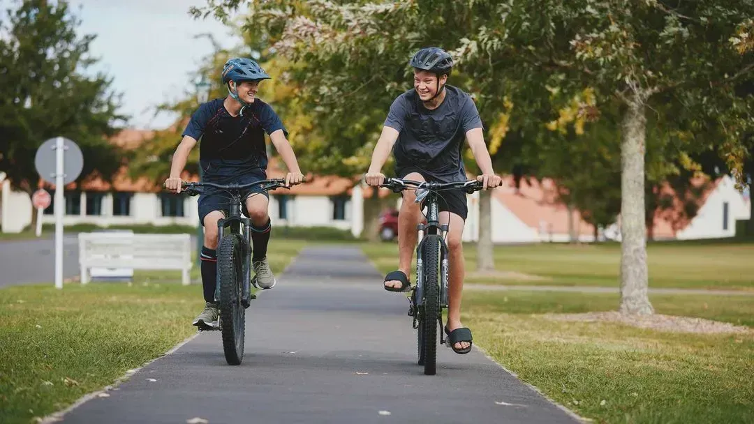 Two students at St. Peter's School enjoy cycling together on a picturesque campus pathway. Surrounded by trees, they embody the vibrant spirit and active lifestyle fostered at this independent school, promoting friendship and outdoor fun.