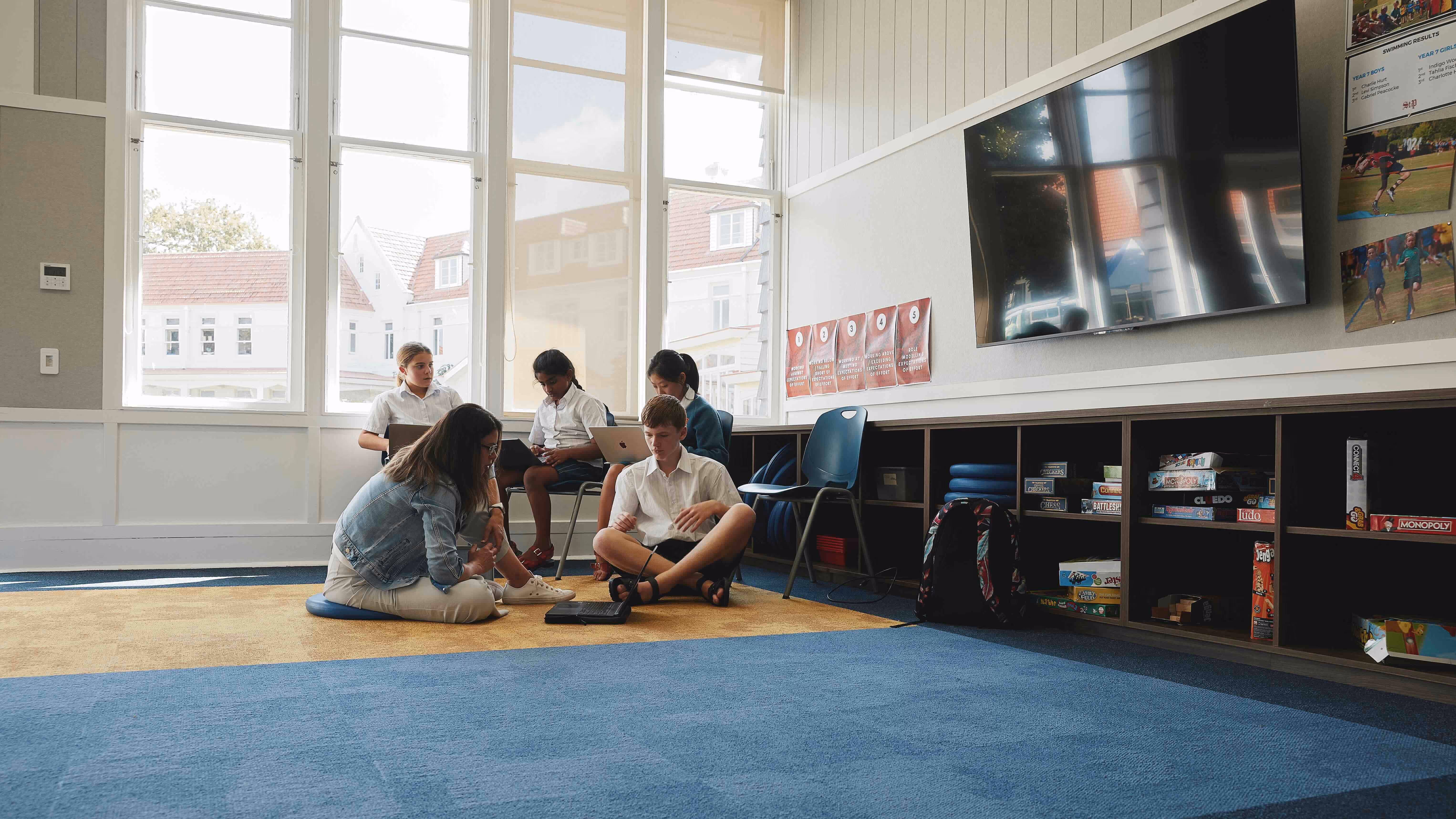 Students collaborating in a modern learning space at St. Peter's School, showcasing academic engagement and teamwork. The bright room features large windows, a television, and a variety of educational resources, promoting a vibrant learning environment.