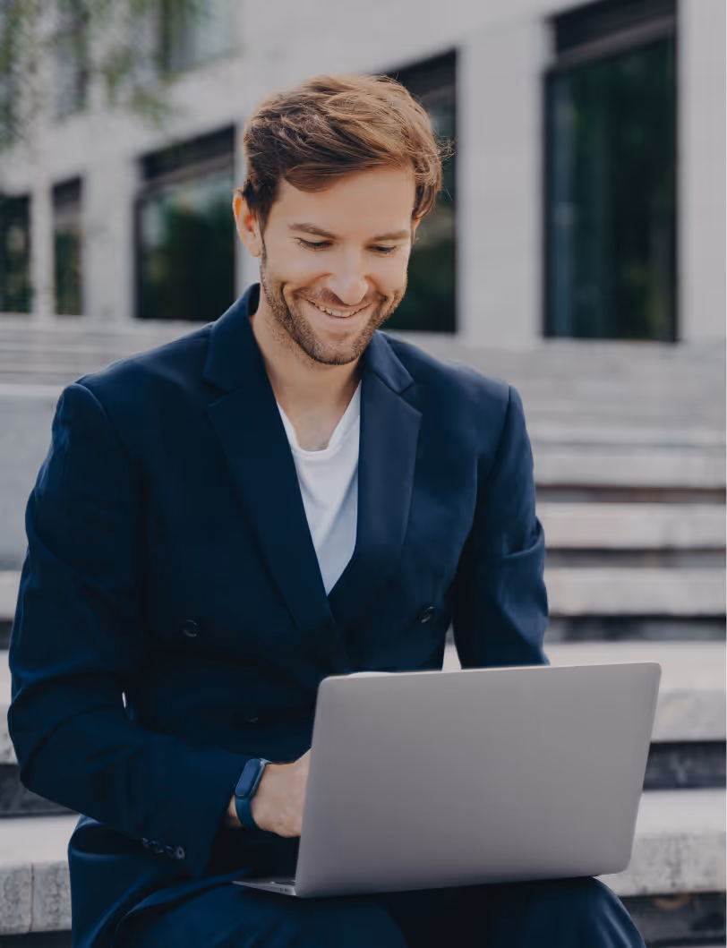 Smiling man in a dark blazer sitting on outdoor steps using a laptop.