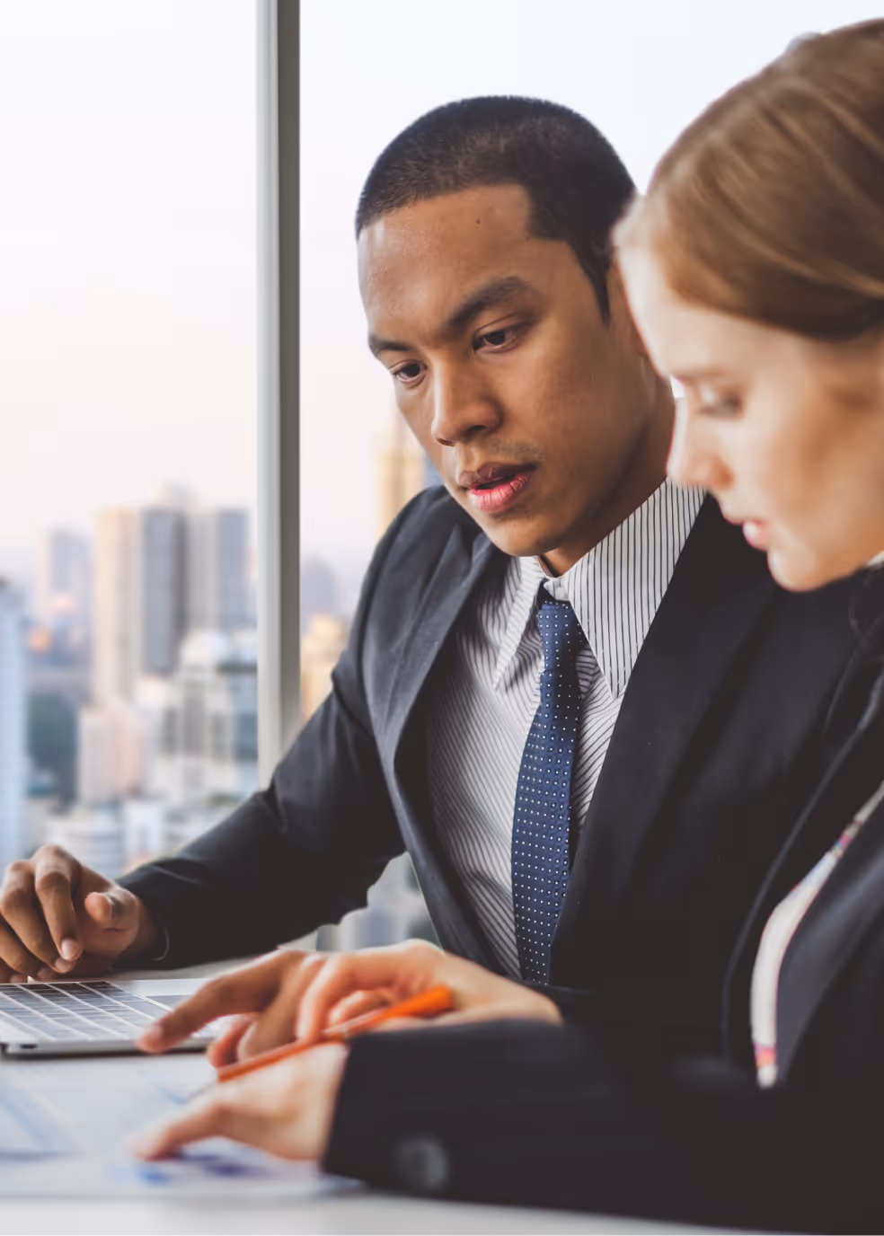 Two business professionals in suits reviewing documents together in an office with a city view.