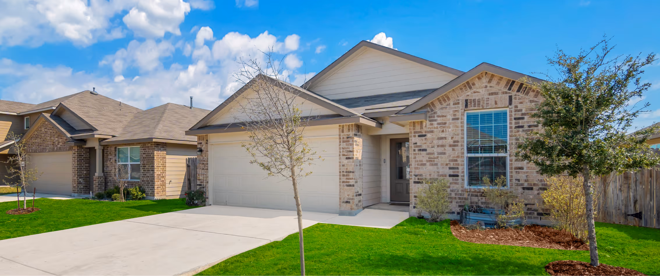 Suburban brick house with a two-car garage, concrete driveway, small front trees, and green lawn under a blue sky.