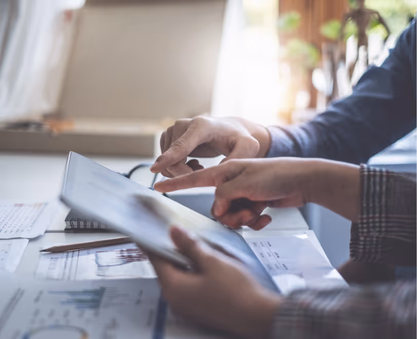 Two people pointing at and interacting with a tablet over a desk with scattered papers and a pencil.