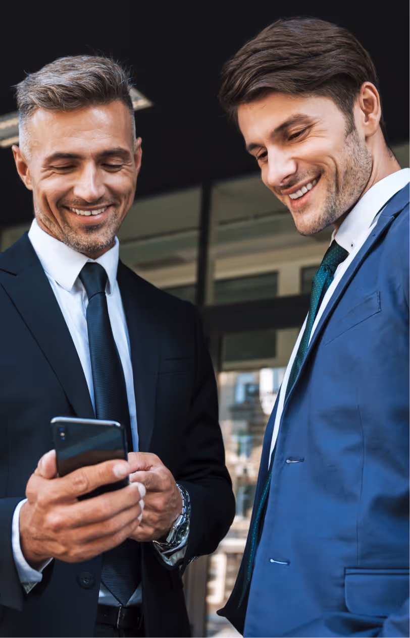 Two businessmen in suits smiling and looking at a smartphone outside a modern office building.