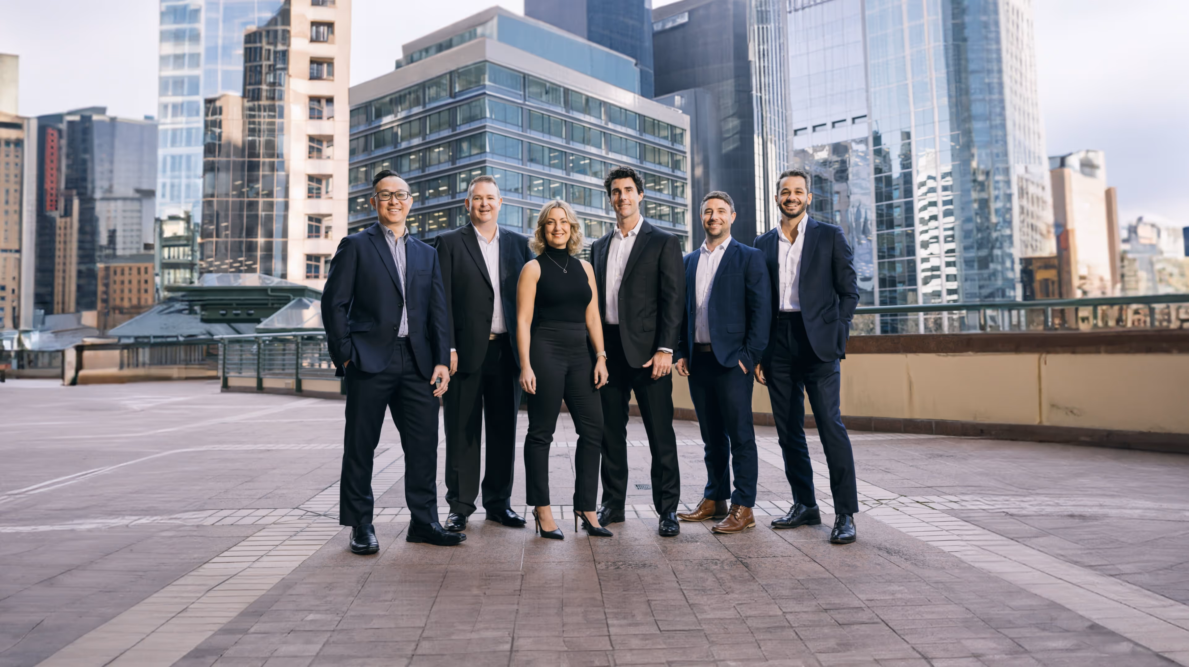 Group of six business professionals standing outdoors on an urban rooftop with tall buildings in the background.