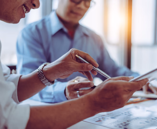 Two people in business attire reviewing documents and a tablet together in a bright office.