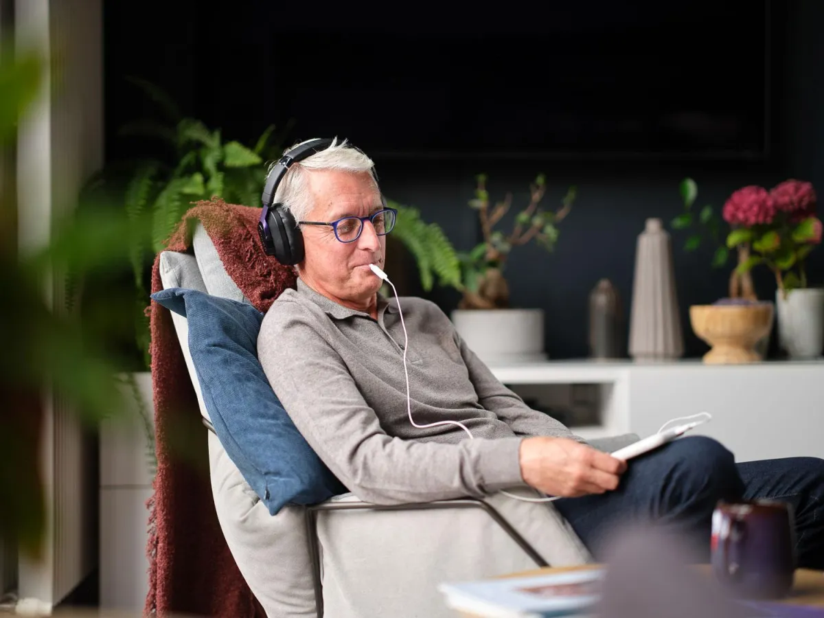 Older man with headphones and glasses sitting in a chair, holding a device with a white wire, surrounded by indoor plants.